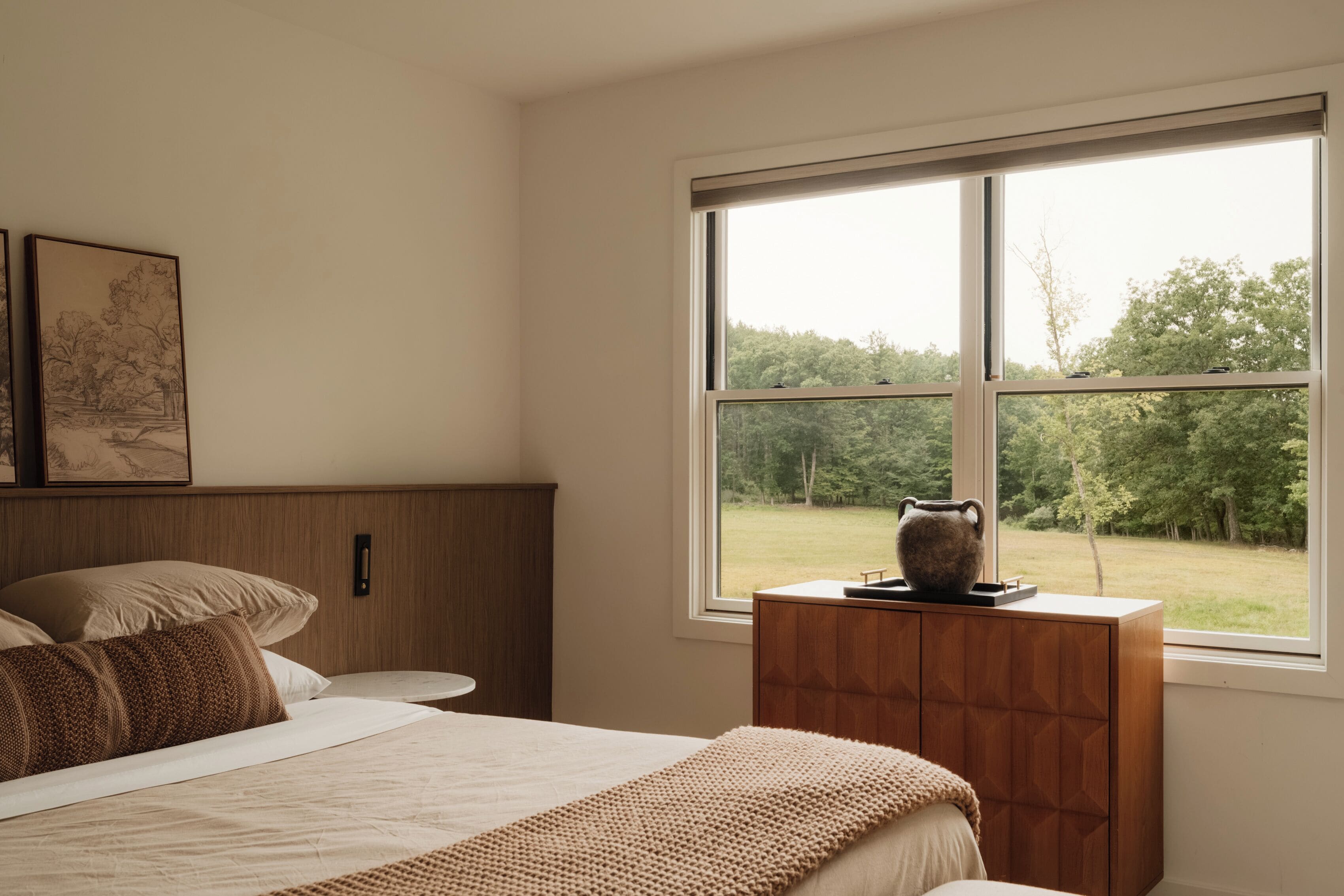 A bedroom with a bed, a nightstand, a window with a view of trees, and a framed picture on the wall.