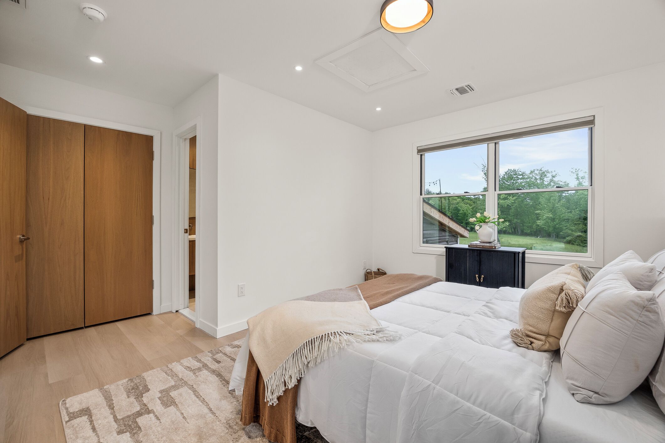 A bedroom with a large bed, a window with a view of greenery, a wooden wardrobe, and a small table with a plant.