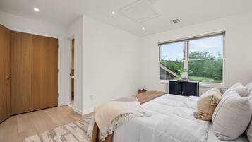 A bedroom with a large bed, a window with a view of greenery, a wooden wardrobe, and a small table with a plant.