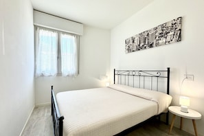 A bedroom with a bed, a window with curtains, a wall-mounted cityscape picture, and a bedside table with a lamp.