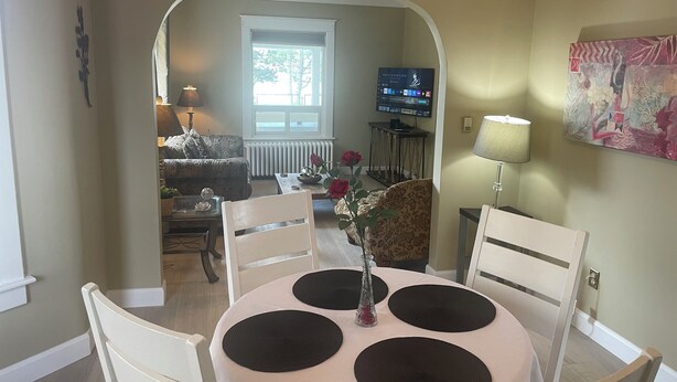 Uma sala de jantar com uma mesa redonda, cadeiras brancas e um vaso com flores.