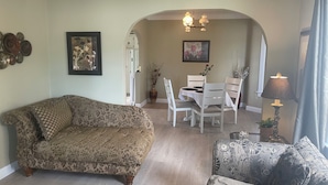 Uma sala de estar com um sofá estampado, uma mesa de centro e uma poltrona estampada. Há uma mesa de jantar com cadeiras e um candelabro ao fundo.