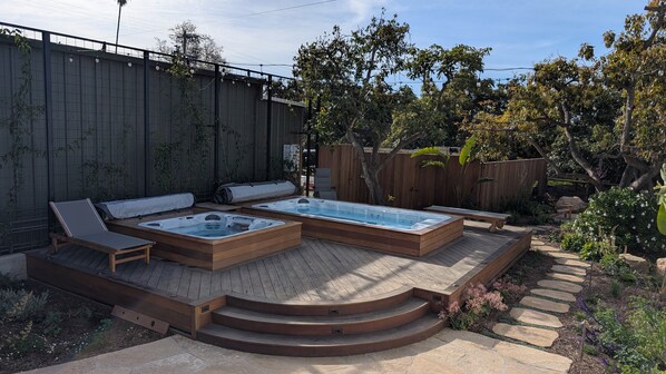 A hot tub with wooden steps and lounge chairs, surrounded by a garden with a wooden fence and trees.