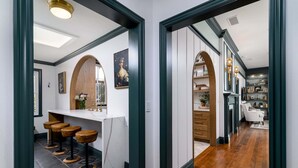 A hallway with a kitchen area, bar stools, and a bookshelf. - Beautiful Magical Historic Webster House at Eleison Farms - Avocado Ranch 3 BD/2 BA (Carpinteria)