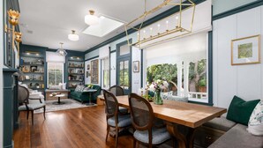 A dining area with a wooden table, four chairs, a sofa, and a bookshelf filled with books and decorative items. - Beautiful Magical Historic Webster House at Eleison Farms - Avocado Ranch 3 BD/2 BA (Carpinteria)