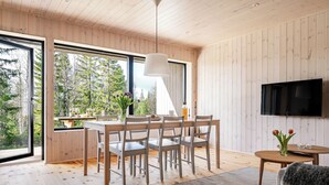 Een moderne eetruimte met een houten tafel en stoelen, een aan de muur bevestigde televisie, en grote ramen met uitzicht op bomen.