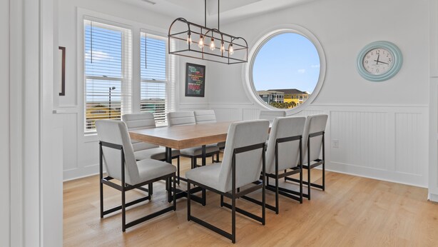A dining area with a wooden table and six chairs, large windows with blinds, a round mirror, and a wall clock. - "Holden Memories" is a 5 Bedroom Second Row Home w/ PRIVATE Pool! (Holden Beach)
