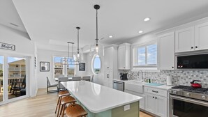A modern kitchen with white cabinetry, a large island with stools, a stove, and a view to the outside. - "Holden Memories" is a 5 Bedroom Second Row Home w/ PRIVATE Pool! (Holden Beach)