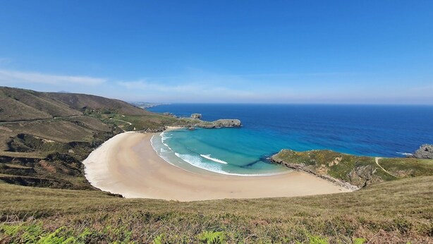 Un paisaje costero con una playa arenosa curva, afloramientos rocosos y un cielo azul despejado.