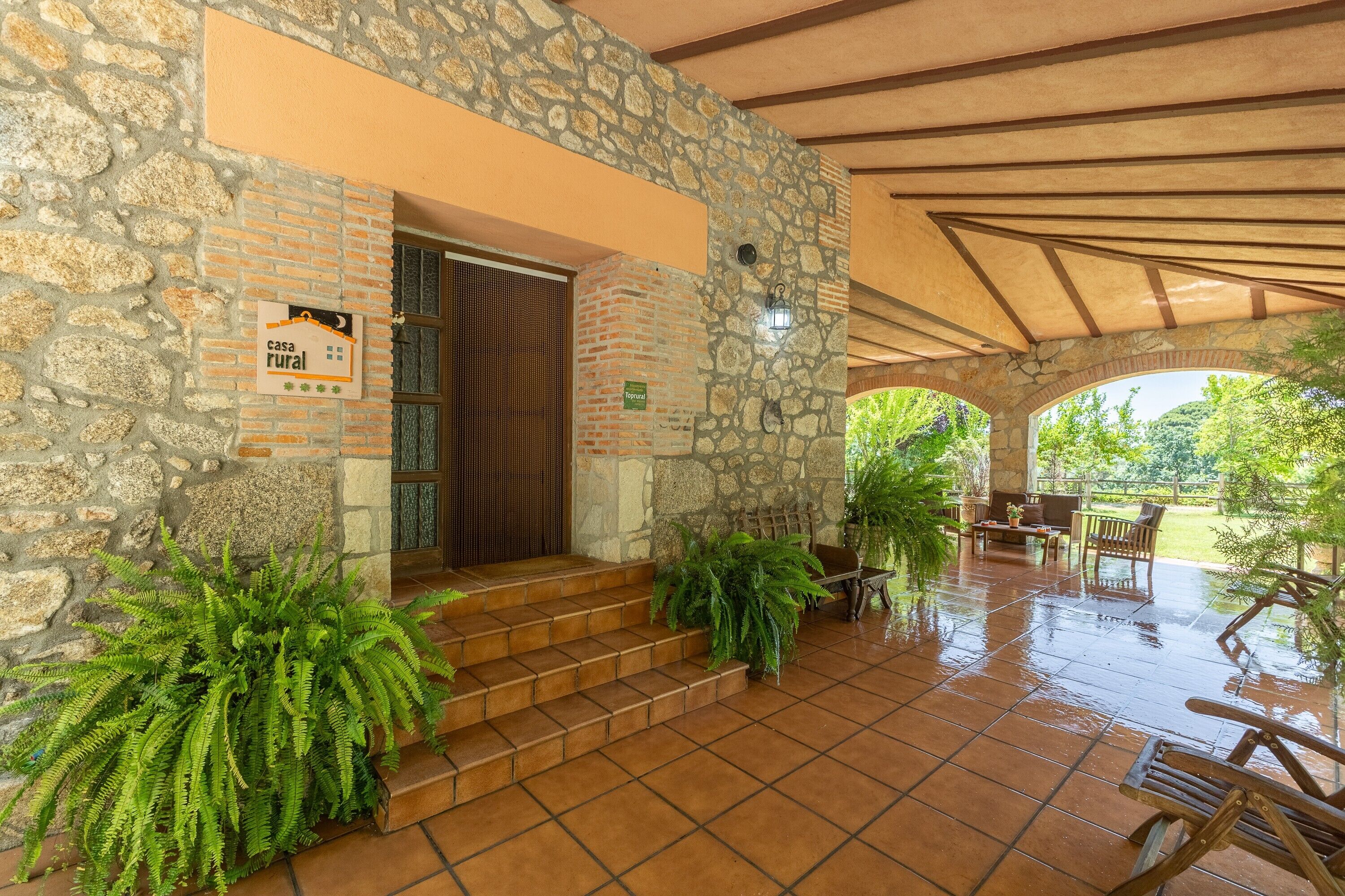 A rustic stone building with a sign indicating it's a rural house, a wooden door, steps leading to a covered patio with outdoor furniture, and potted plants.