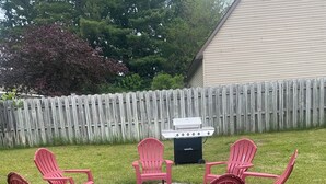 Ein Garten mit Feuerschale, vier Adirondack-Stühlen und einem Grill.