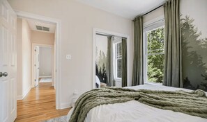A bedroom with a large bed, a view of trees through the window, and a door leading to another room. - Hot Tub-Movie Rm-Game Rm (Asheville)