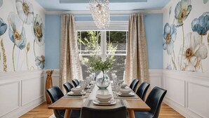 A dining room with a wooden table set for six, flanked by blue floral wallpaper, a large window with curtains, and a chandelier. - Hot Tub-Movie Rm-Game Rm (Asheville)