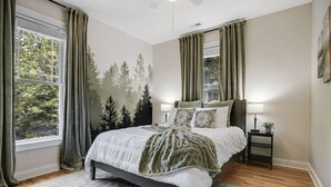 A bedroom with a bed, two bedside tables, a window with curtains, and a wall mural. - Hot Tub-Movie Rm-Game Rm (Asheville)