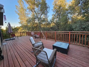 Eine Holzterrasse mit Korbstühlen und einem Tisch, umgeben von einem Hollerleger und Bäumen.
