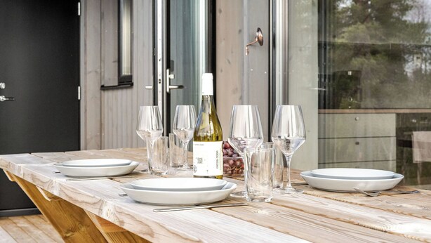 A dining table set with plates, wine glasses, and a bottle of wine, positioned in front of a wooden door and large windows. - Renstråket 1001 by Interhome (Tännäskröket)