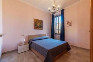 A bedroom with a bed, nightstand, lamp, artwork, window with curtains, and a chandelier.