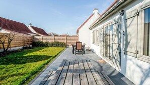 Una terraza de madera con mesa y sillas, rodeada por un edificio blanco con ventanas grandes y techo de tejas rojas.