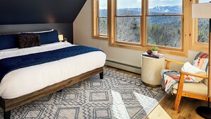 Une chambre avec un grand lit, une vue sur les montagnes par la fenêtre, une chaise et une petite table sur laquelle est posée une plante.