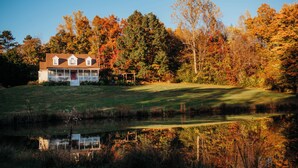Une maison avec un porche, un étang au premier plan et des arbres aux feuilles d’automne en arrière-plan.