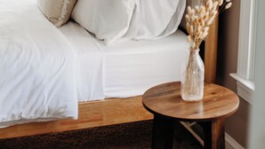 Un lit bien fait, avec du linge de lit blanc, et un vase de fleurs séchées posé sur une petite table en bois à côté.
