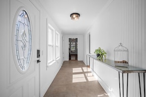Un couloir aux murs blancs, doté d’un plancher en bois, d’une porte en verre ornée d’un motif décoratif, d’un luminaire et d’une petite table sur laquelle sont posés une plante et une cage à oiseaux.