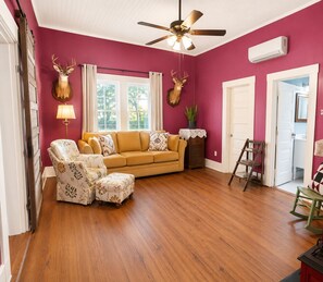 Una habitación con piso de madera, un sofá color beige, un sillón estampado, un ventilador de techo y dos decoraciones de cabeza de ciervo en la pared.