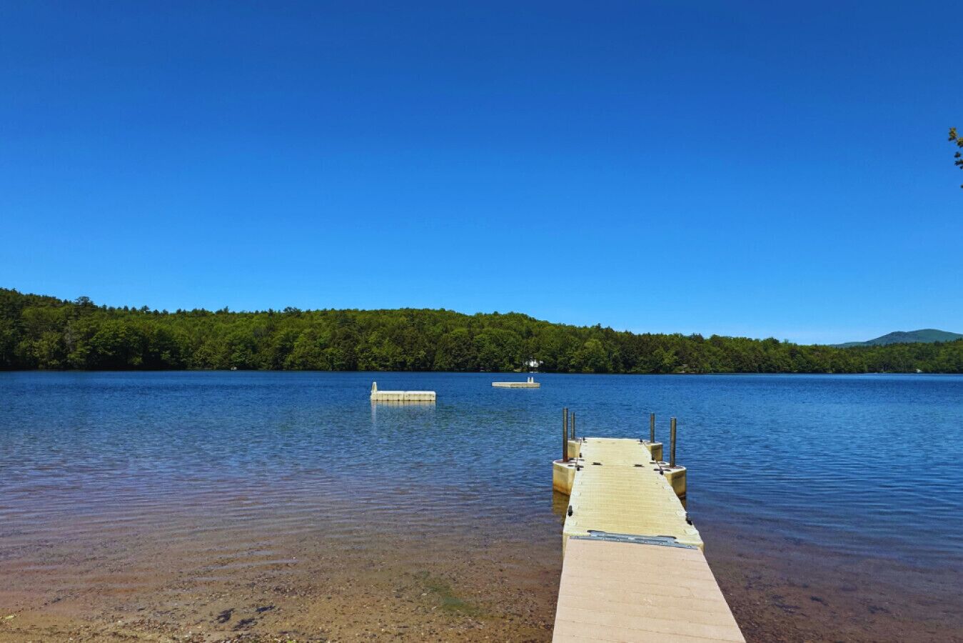 Une jetée en bois s’étendant sur un lac paisible, bordée d’une rive boisée et surplombée d’un ciel bleu clair.