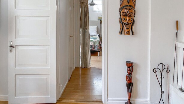 Un couloir avec un plancher en bois, une porte blanche et, au fond, un lit recouvert d’une courtepointe à motifs floraux.