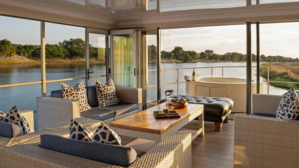 A modern, open-plan living area with a large glass window overlooking a river, furnished with wicker sofas and a wooden coffee table. - Chobe Princesses By Mantis (Kasika)