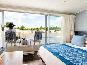 Habitación de hotel con un ventanal grande que ofrece vistas de la fauna y un río. Hay una cama con colcha azul estampada, dos sillones con mantas y una mesa con una bebida y una cámara.