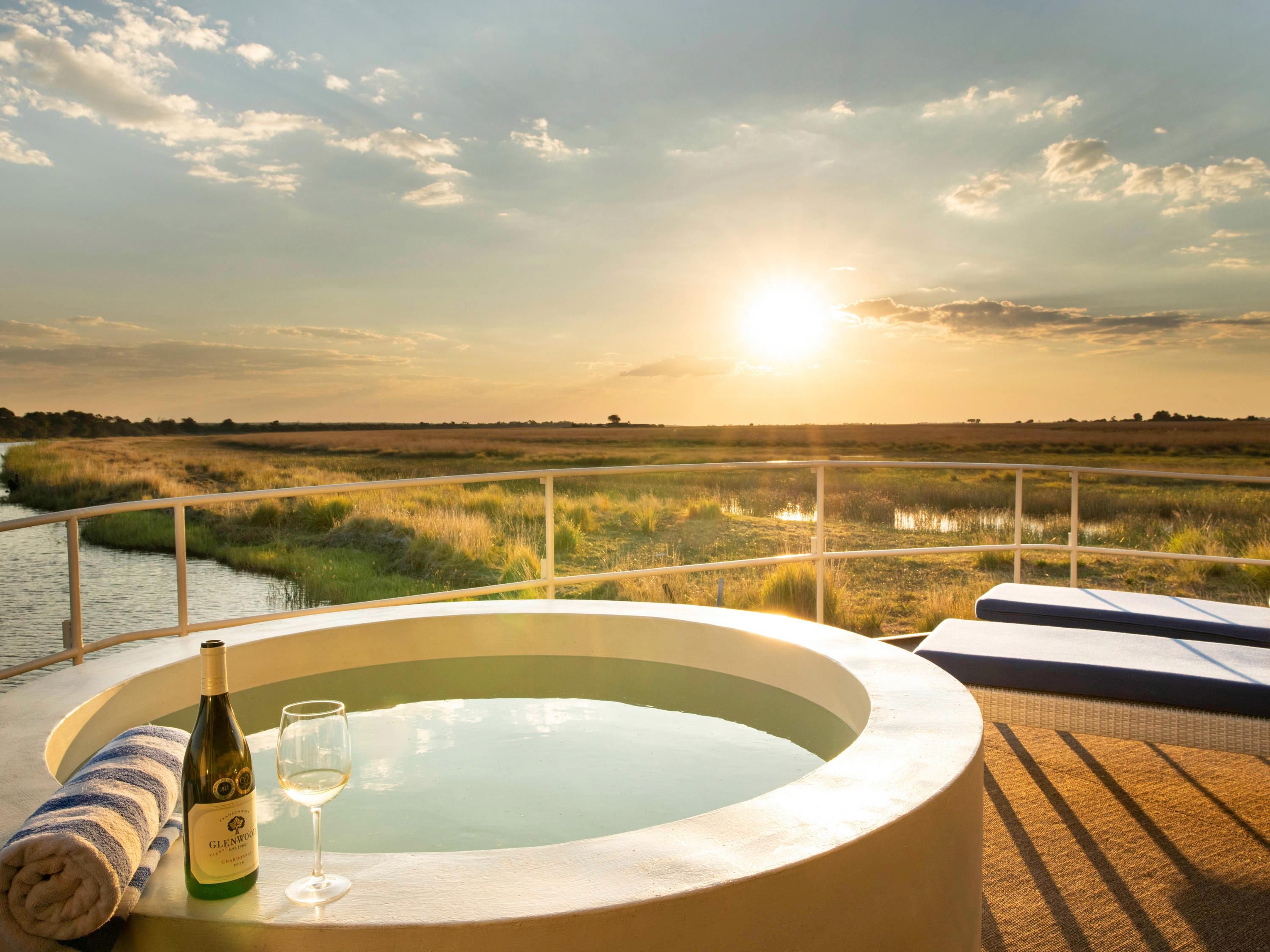 A hot tub with a bottle of Glenfiddich whisky and a glass, overlooking a marshy landscape with a sunset in the background.