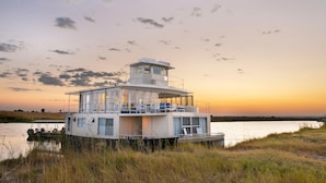 A houseboat with a covered deck and multiple levels, situated on a body of water with a sunset in the background. - Chobe Princesses By Mantis (Kasika)