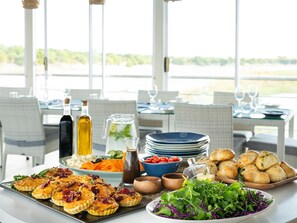 A dining table set with various dishes, including quiches, salads, and bread, in a bright room with large windows. - Chobe Princesses By Mantis (Kasika)