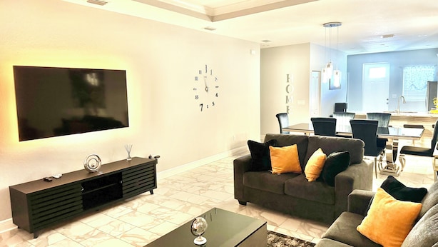 A modern living room with a flat-screen TV, a dark sofa set, a coffee table, and a dining area in the background.