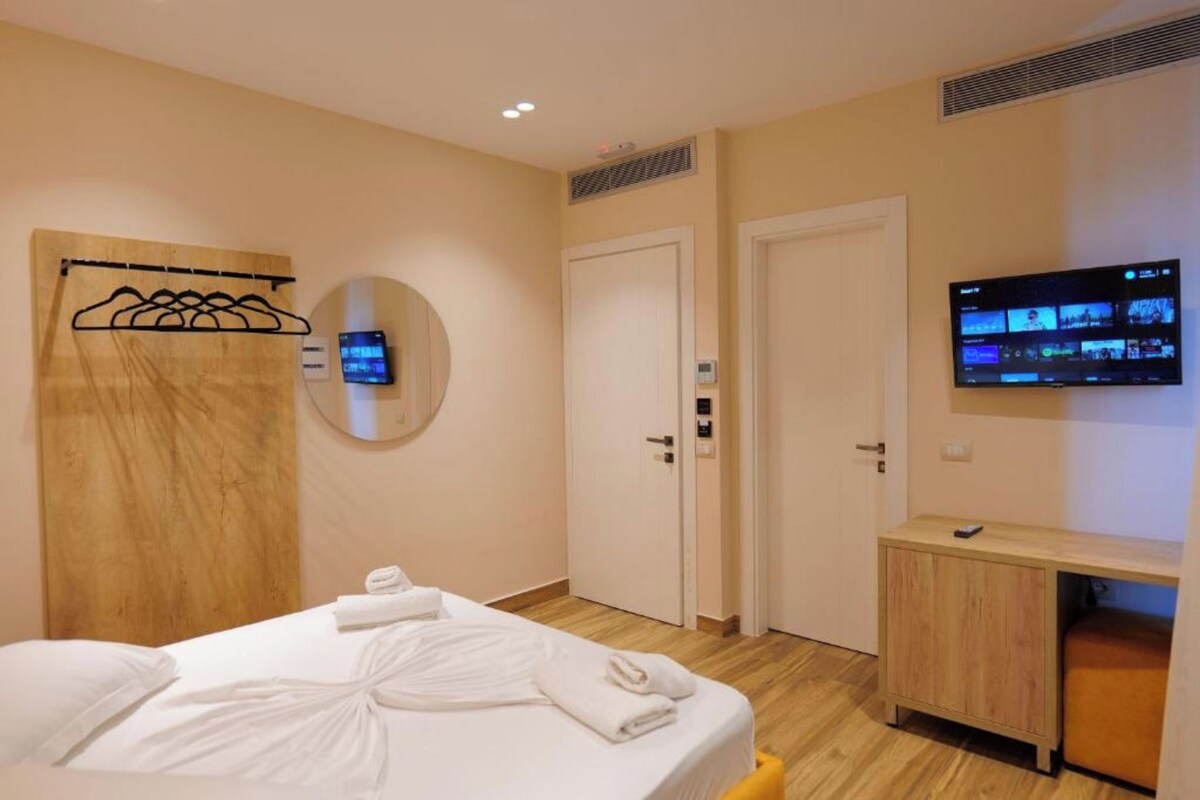 Quarto de hotel com cama, televisão, espelho e cabeceira de madeira.