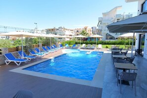A swimming pool with sun loungers and umbrellas, surrounded by outdoor seating and a view of residential buildings. - Hotel Alexander (Sarandë)