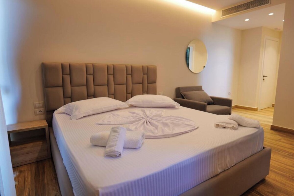 Cama bem arrumada com cabeceira estofada, espelho redondo na parede e piso de madeira.