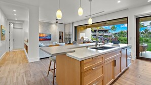 Uma cozinha moderna com uma ilha grande, iluminação pendente e vista para uma área externa tropical.
