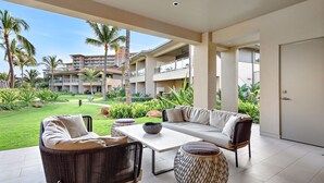Uma área de estar ampla ao ar livre, com sofá, cadeiras e mesa, com vista para um jardim bem cuidado e um edifício alto ao fundo.