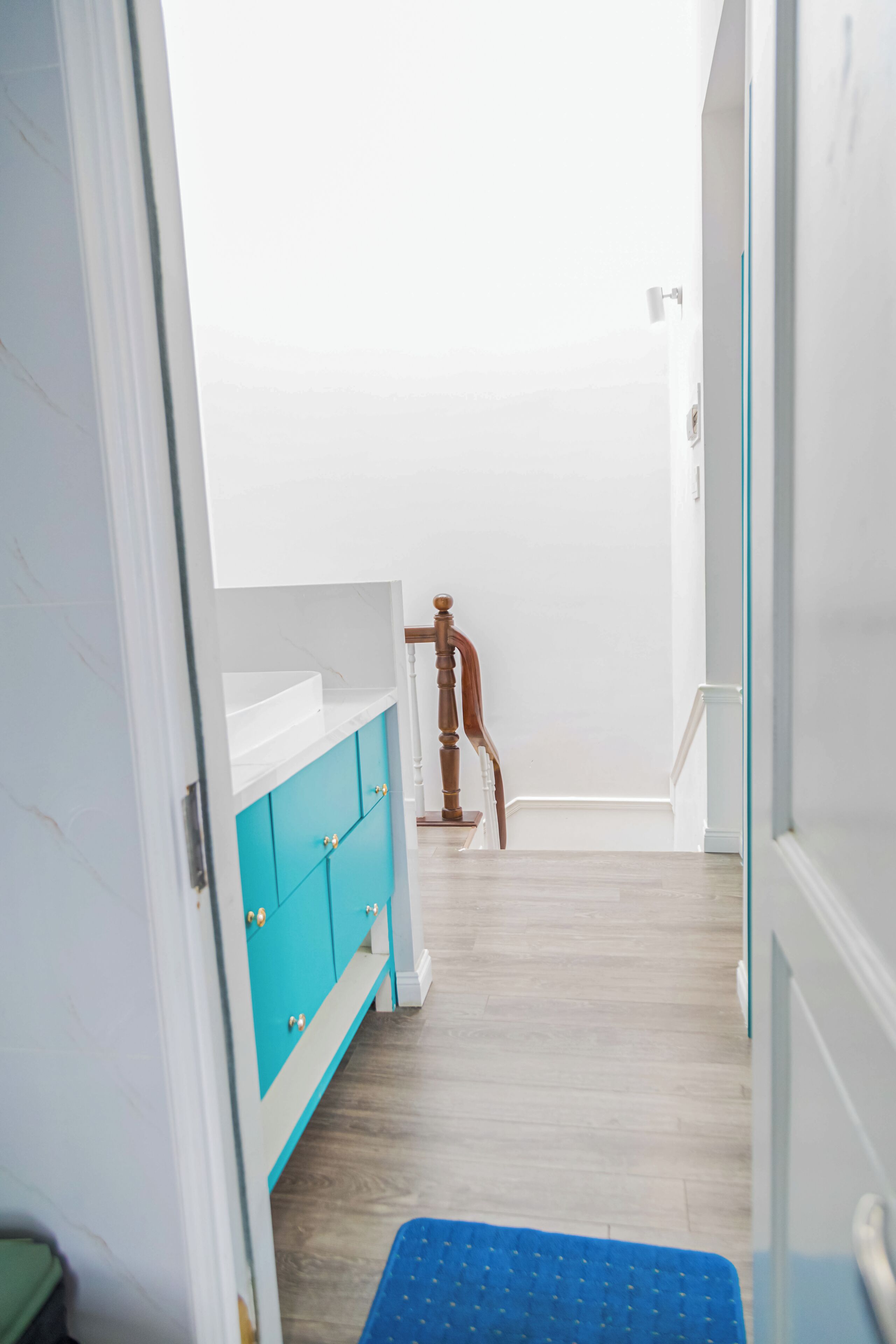 A narrow hallway with a wooden door, a blue mat, and a white cabinet with teal doors.