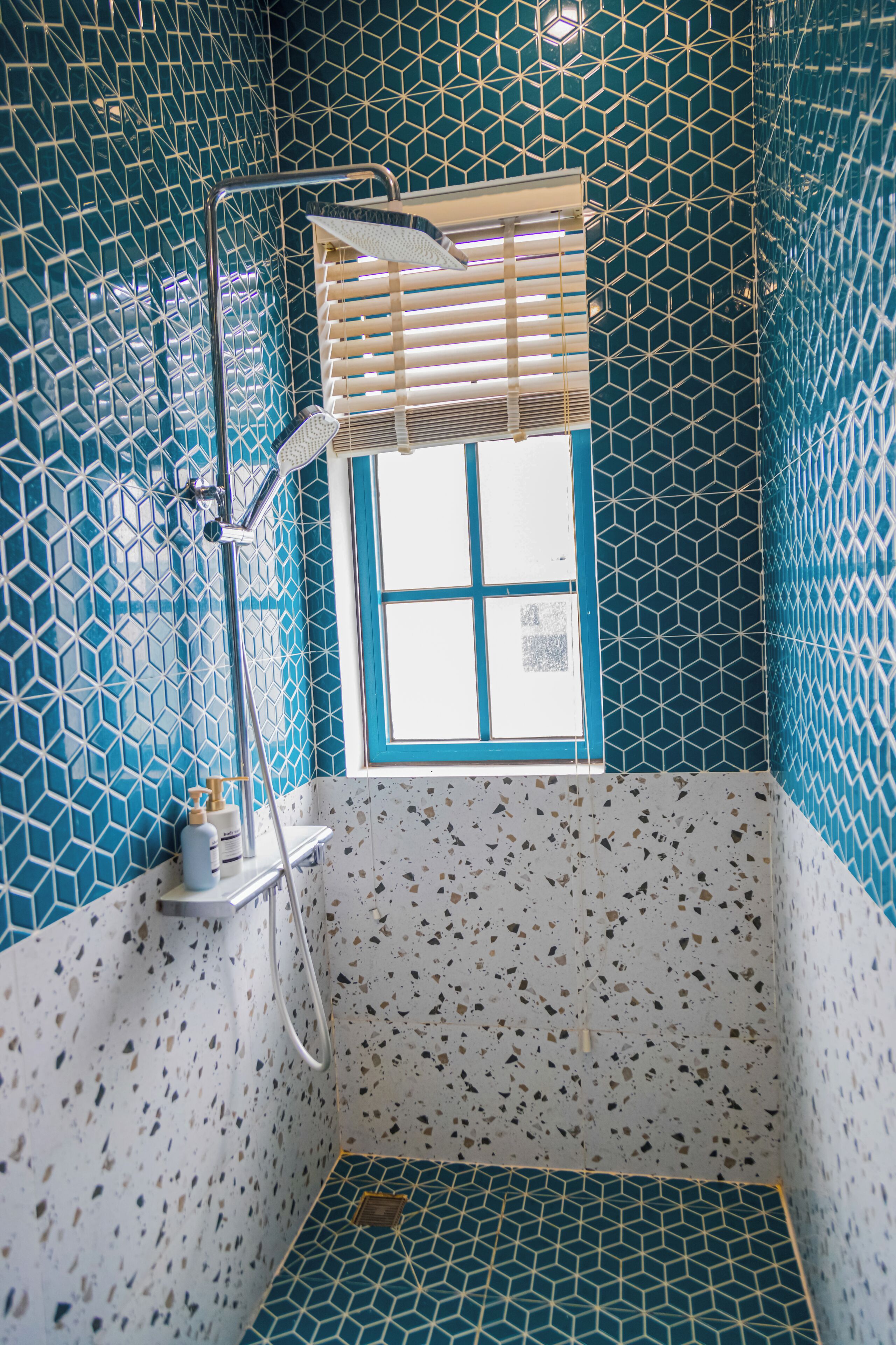 A bathroom with a geometric tile pattern, a window with blinds, and a shower area.