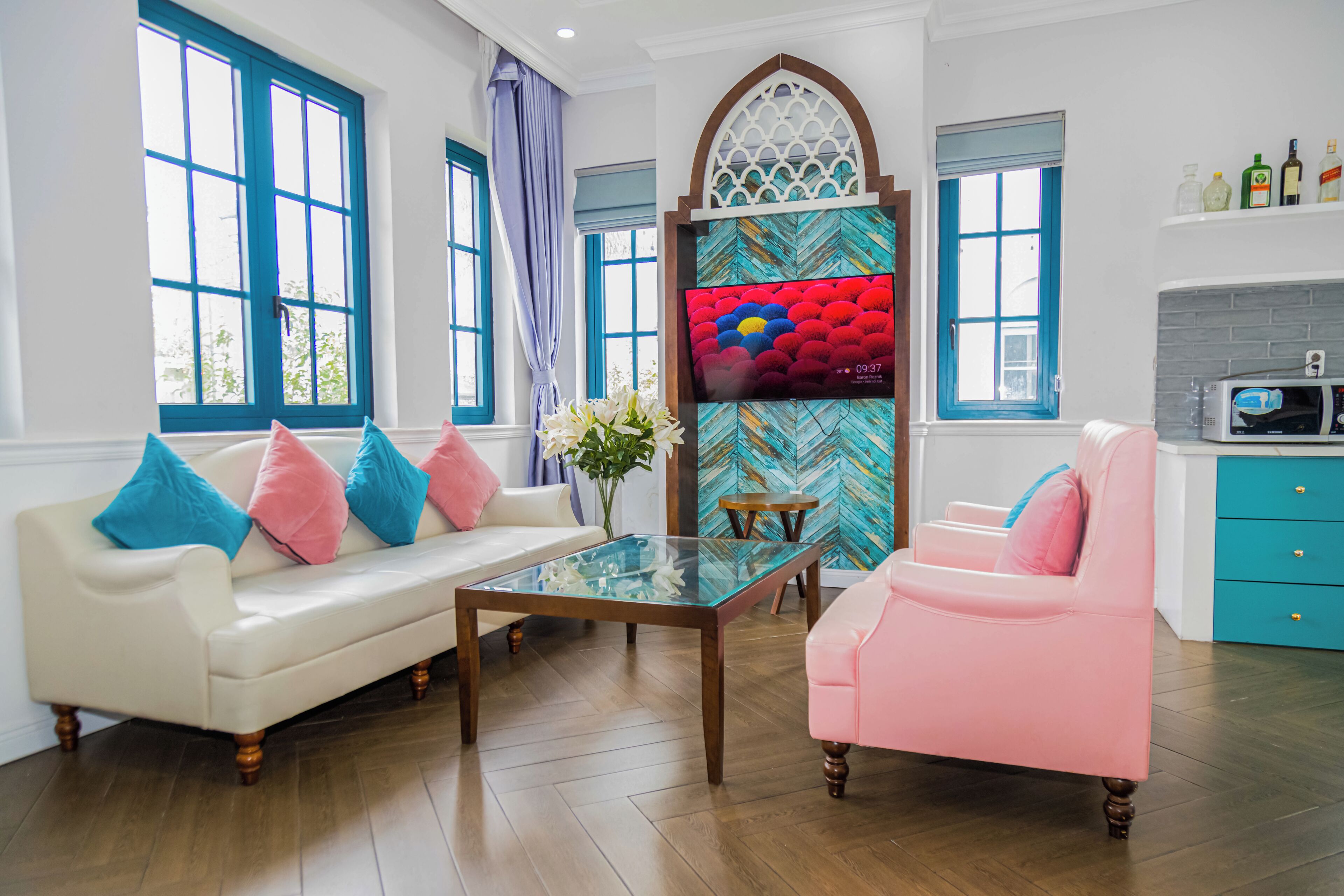 A living room with a white sofa, a pink armchair, a glass coffee table, and a TV mounted on a wall with a decorative screen.