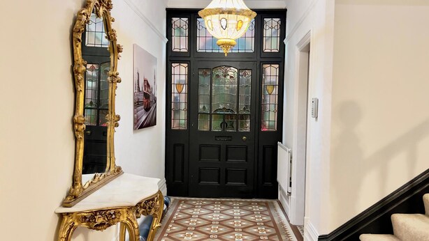 Un pasillo con una puerta negra, un espejo con marco dorado, una lámpara de araña y un suelo con diseño.