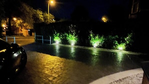 Scena notturna di una strada con un'auto parcheggiata a sinistra, un marciapiede pavimentato e piante illuminate di verde lungo il cordolo.