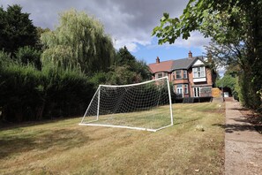 Una portería de fútbol en un césped, con una casa de ladrillo al fondo.