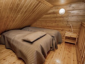 A wooden cabin room with a sloped ceiling, two beds with matching bedding, a bedside table, and a lamp.