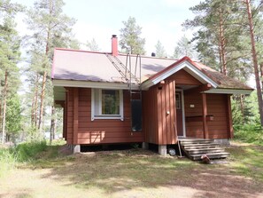 A small, single-story cabin with a red roof, a chimney, and a small porch with steps leading up to the entrance.