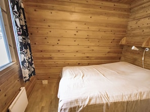 A small, single-bed room with wooden walls, a window with curtains, and a bedside lamp.