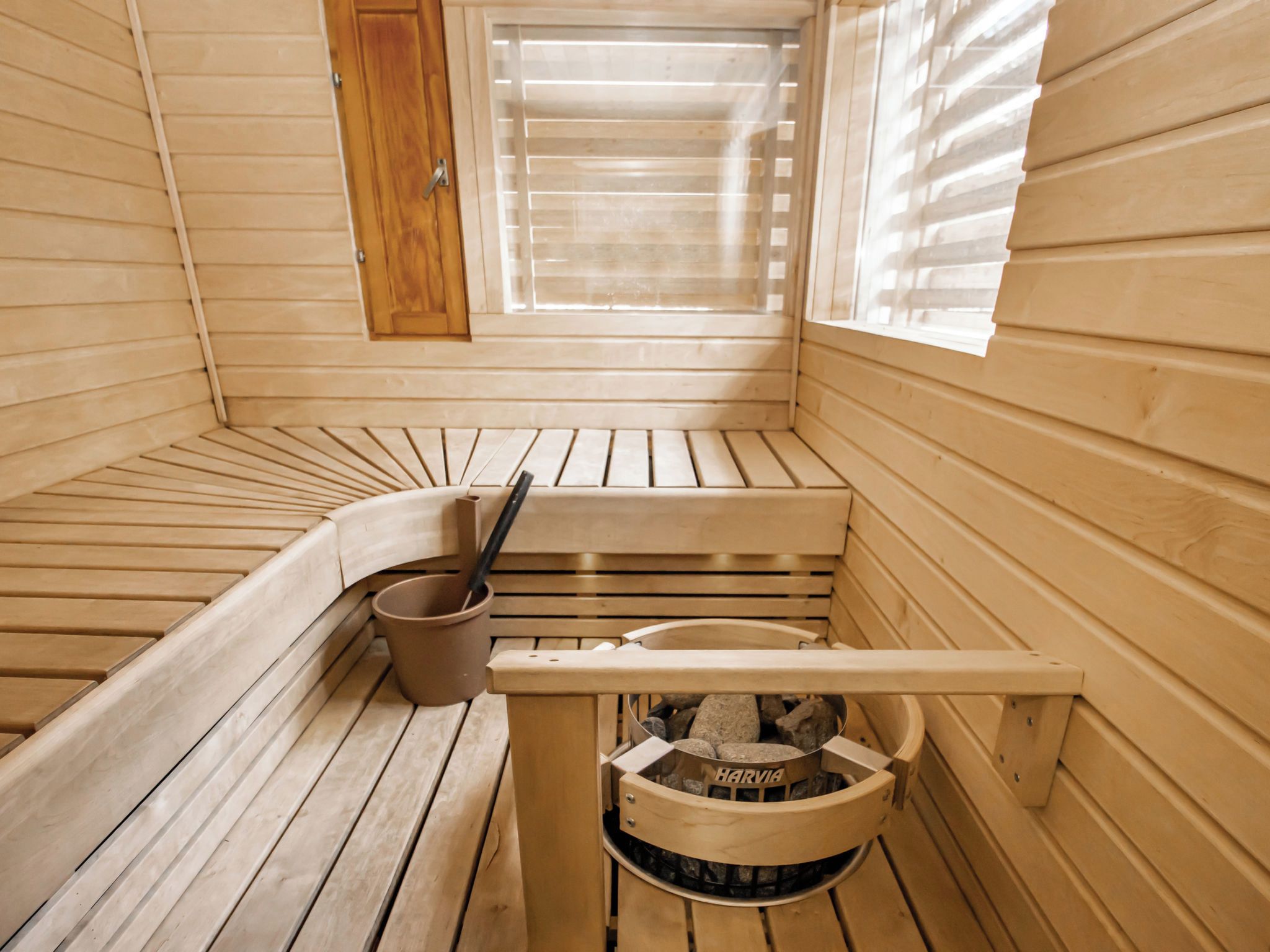 Una sala de sauna con bancos de madera, un cubo y un recipiente lleno de piedras con la etiqueta 'HARYA'.